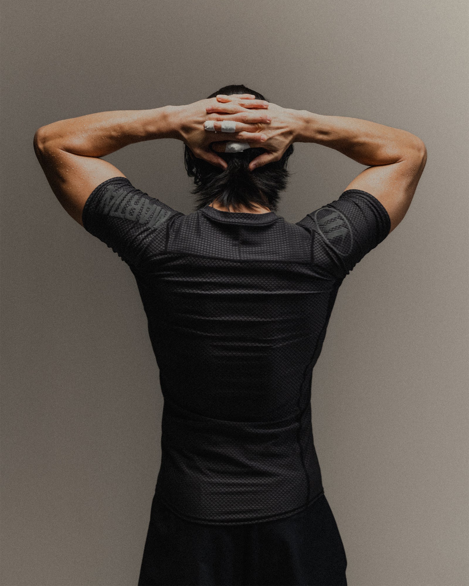 Man wearing black fight shorts and black Martial rashguard on a gray background, standing to the back to show the shoulder branding.