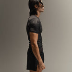 Man wearing black fight shorts and black Martial rashguard on a gray background, standing to the side profile.