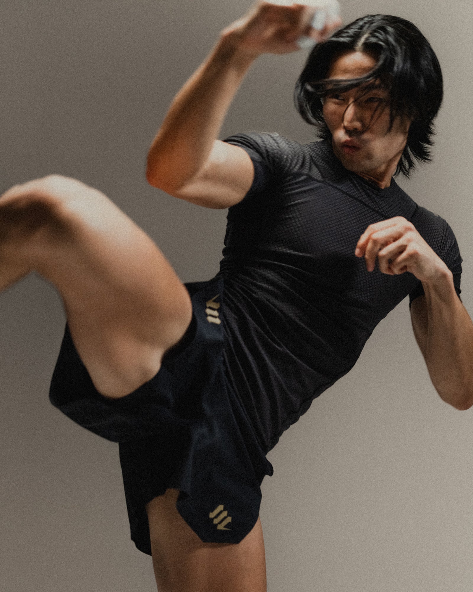 Man in black Martial rashguard and fight shorts performing a high kick on a neutral background.