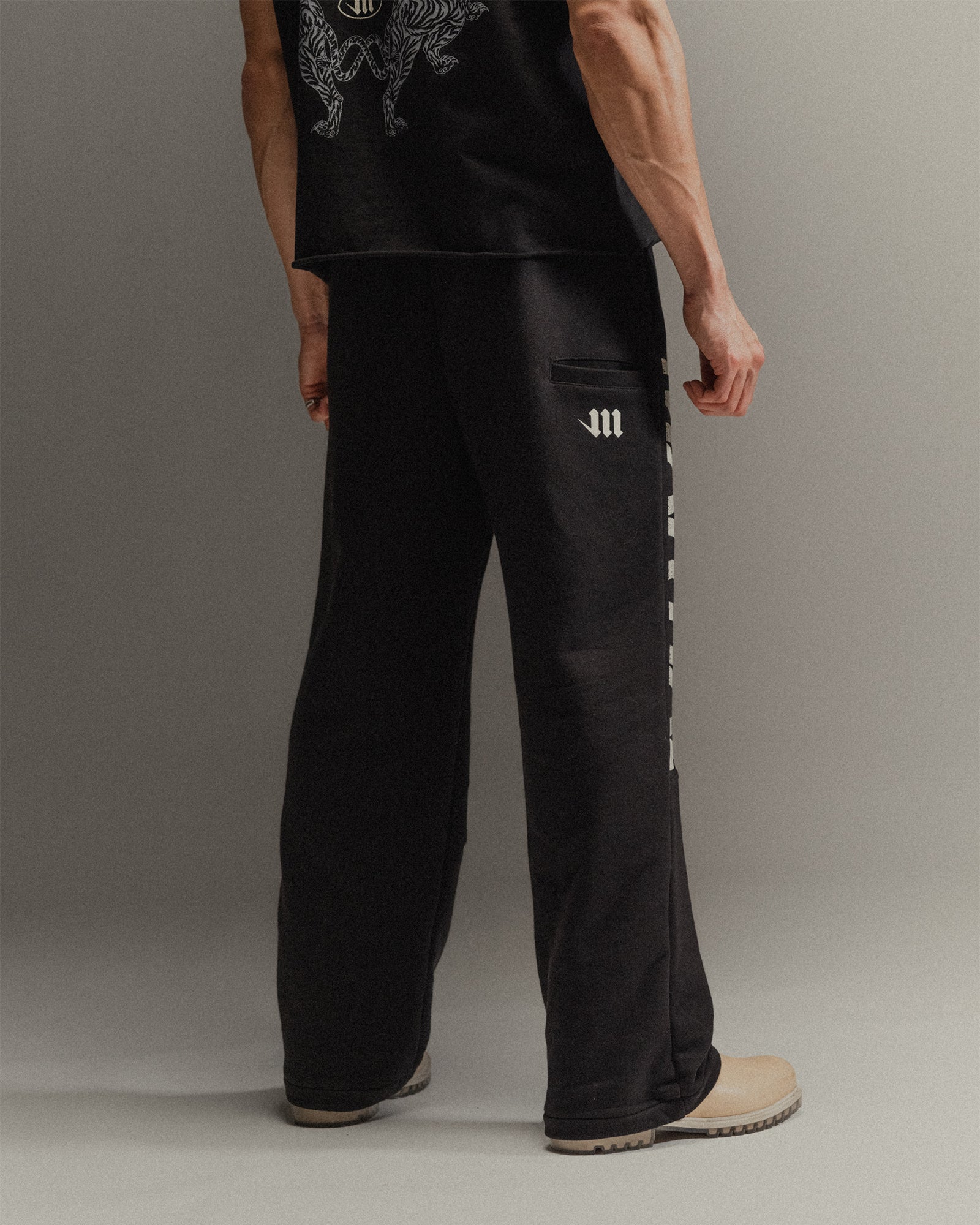 Man wearing black pants with a side drop phone pocket and cream 'M' logo on a gray background