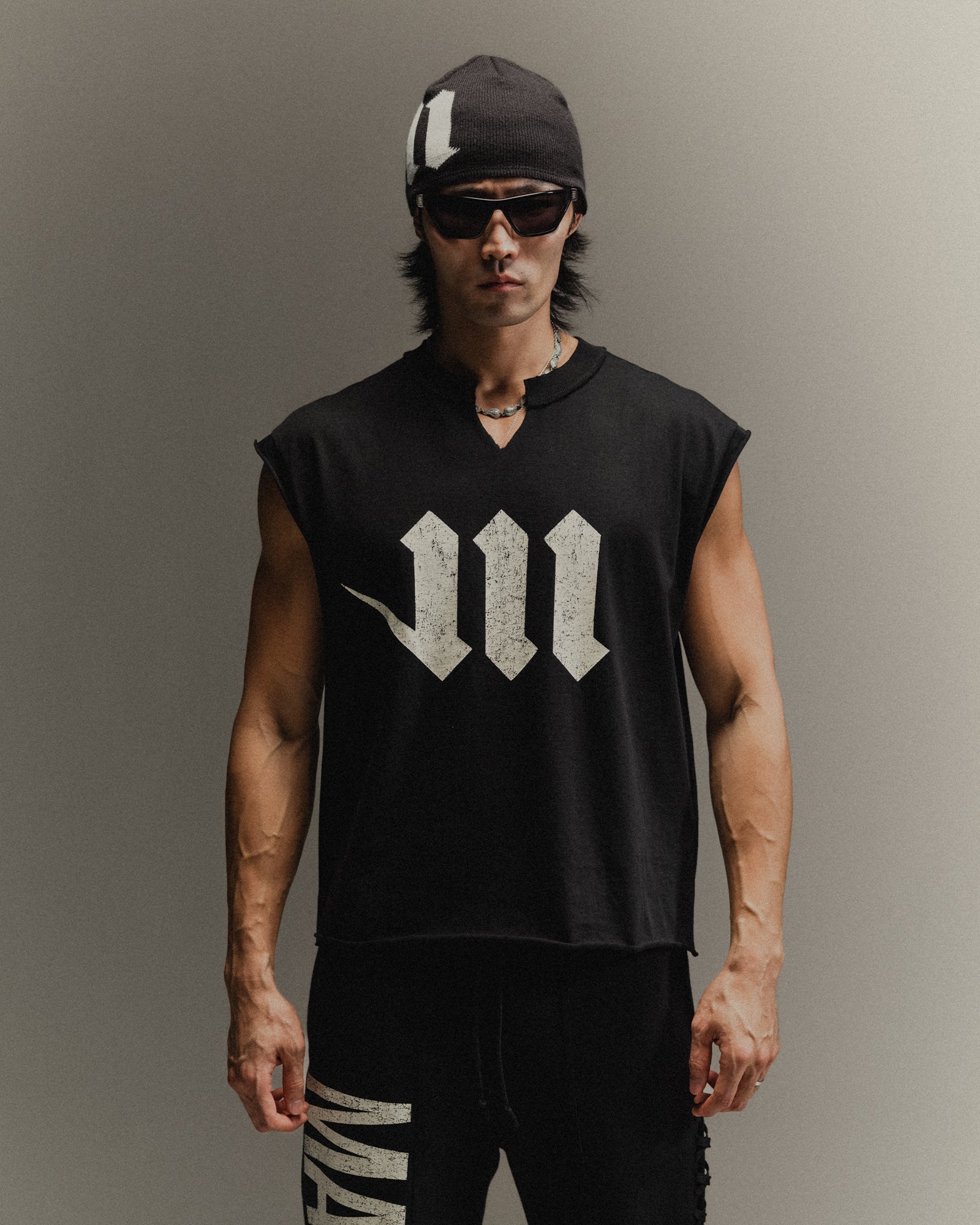 Man wearing black skullcap, sunglasses, black Martial cut-off shirt with a neck slit and cream logos, and black Martial sweats on a neutral backdrop.