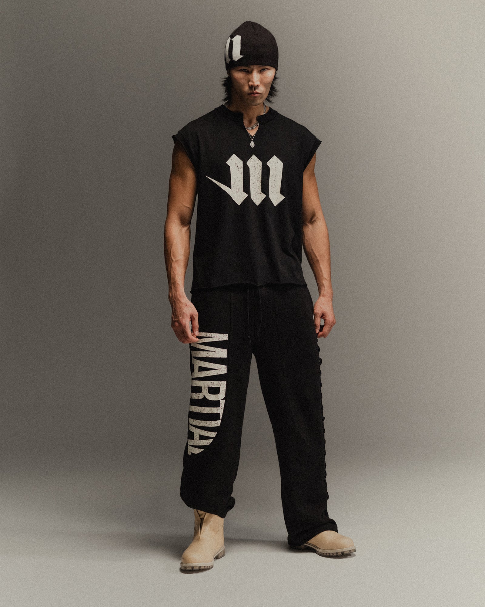 Man wearing a black sleeveless shirt with a white 'M' logo and black pants with 'Martial' printed on them, standing against a gray background.