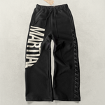 Black sweatpants with 'MARTIAL' text graphic and lace weave design laying on a light gray background