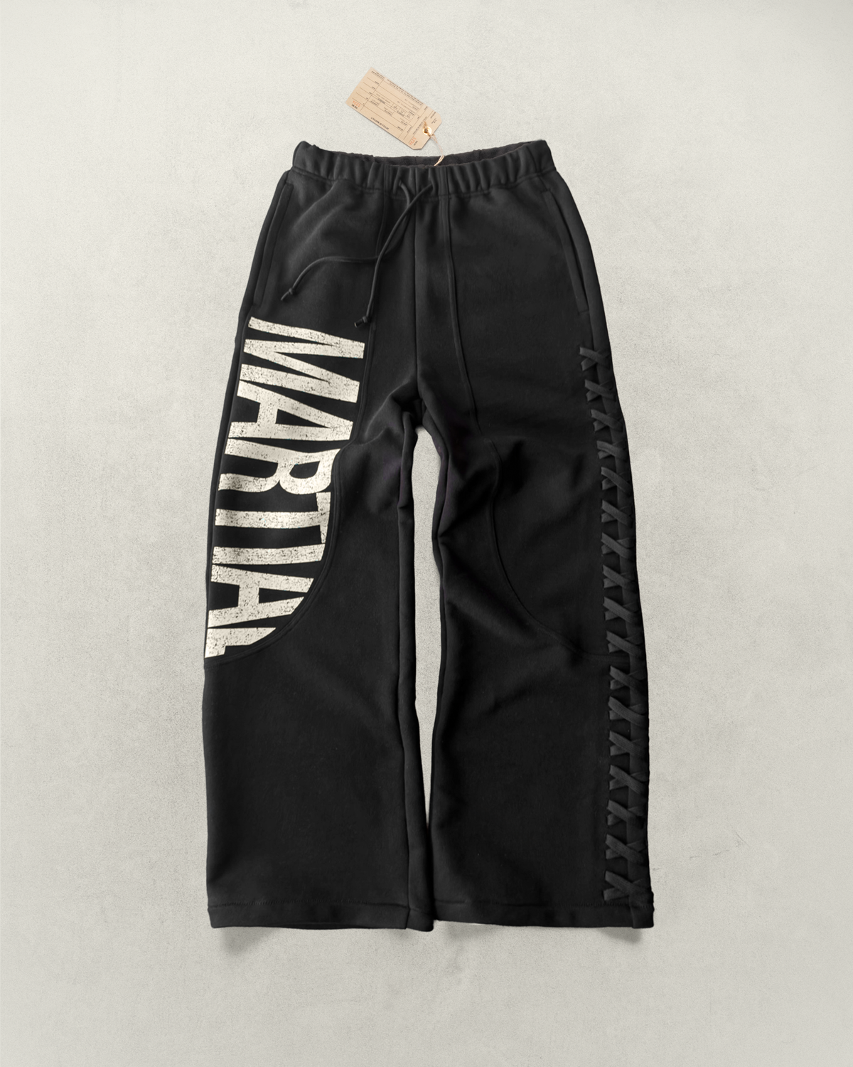 Black sweatpants with 'MARTIAL' text graphic and lace weave design laying on a light gray background