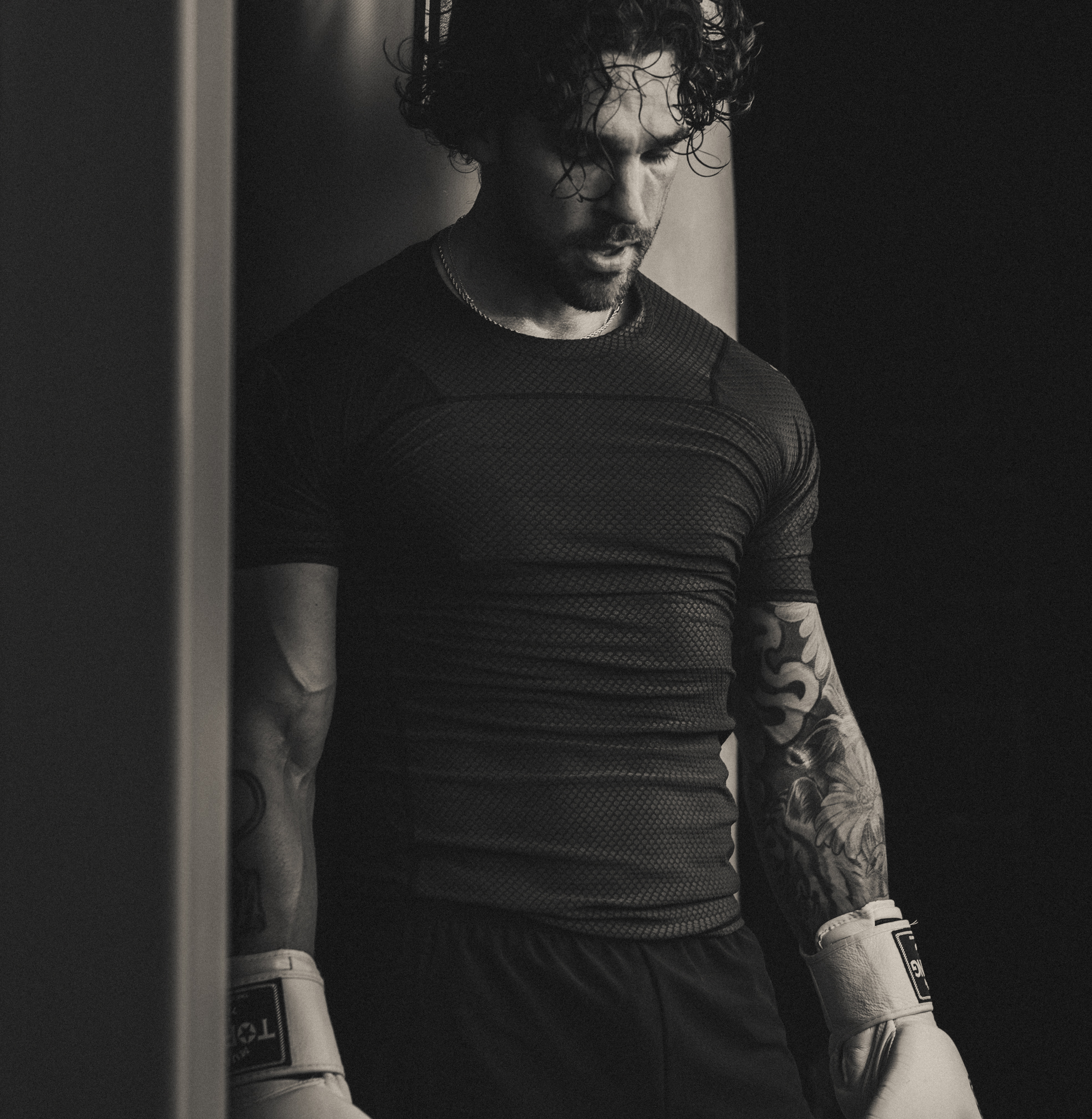 Black and white photo of man resting in boxing gym wearing black rash guard and black fight shorts.